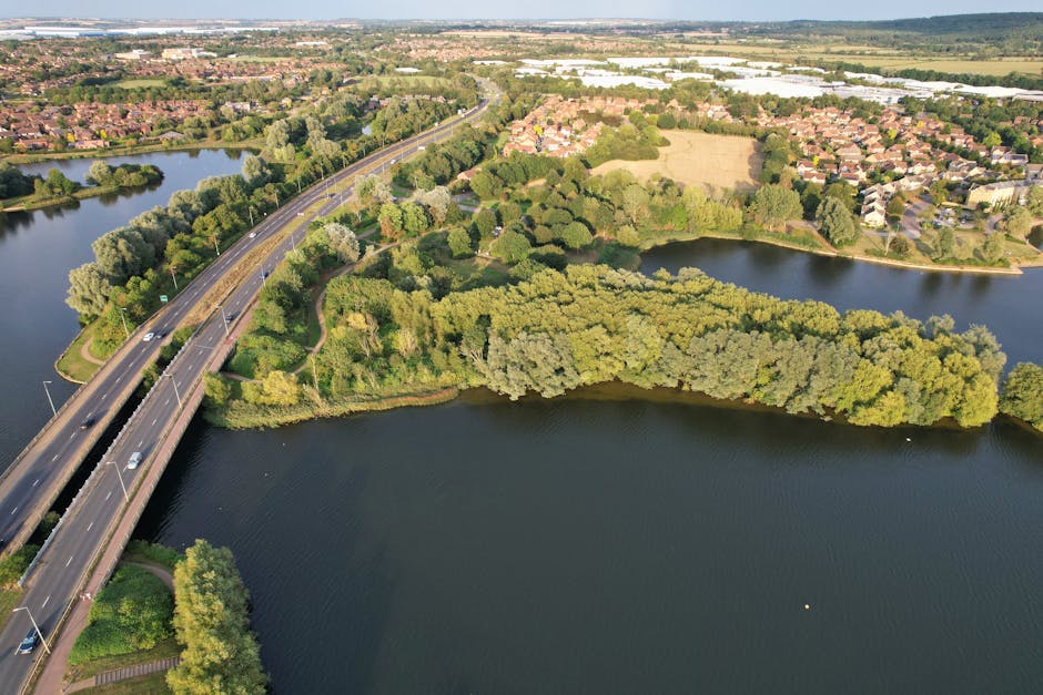 An aerial view of a section of the River Roding winding through a green landscape near Hainault, with a bridge crossing over the waterway. The scene shows dense tree cover along the riverbanks, open grassy areas, and a residential estate comprised of rows of houses with pitched roofs in the distance. The bridge is a multi-lane road used for vehicle transportation, with several cars visible traveling in both directions. The surrounding environment is lush and well-maintained, under bright daylight with clear visibility of the natural and built features. This image illustrates the scenic environment around Hainault, often considered during house removals and relocation planning. Man with Van Hainault offers professional moving services in this area, supporting clients with efficient furniture transport and packing during home relocations.