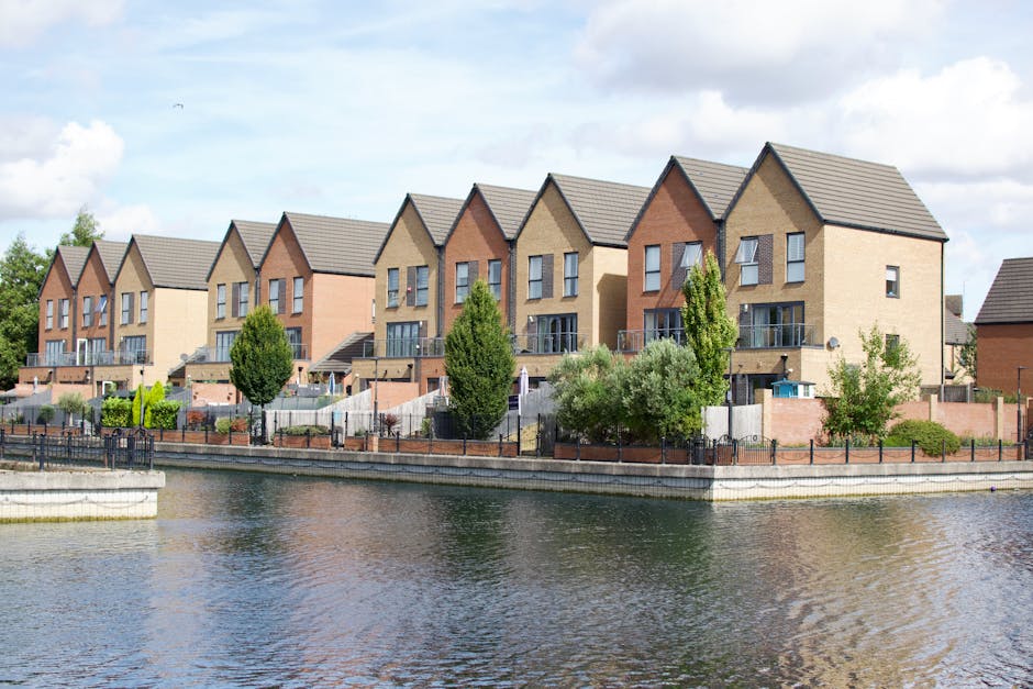 A row of modern, multi-storey houses with pitched roofs and large windows lined along a waterway on a bright, partly cloudy day. Each house has a small balcony or terrace, some with glass railings, and is set behind a low brick wall or fencing. In front of the houses, there are a few trees and shrubs, including tall, slim trees and broader, leafy bushes. The water in the foreground is calm, reflecting the houses and sky, with a paved walkway and a metal railing along the edge. Inside a property boundary, visible elements include cardboard boxes, plastic wrapping, and packing blankets on the pavement, with people engaged in lifting and loading furniture and boxes onto a vehicle. A blue moving van is partially visible, with a ramp and straps securing items for transport, indicating a home relocation or furniture transport process, with the scene typical of house removals carried out by [COMPANY_NAME], Man with Van Hainault.