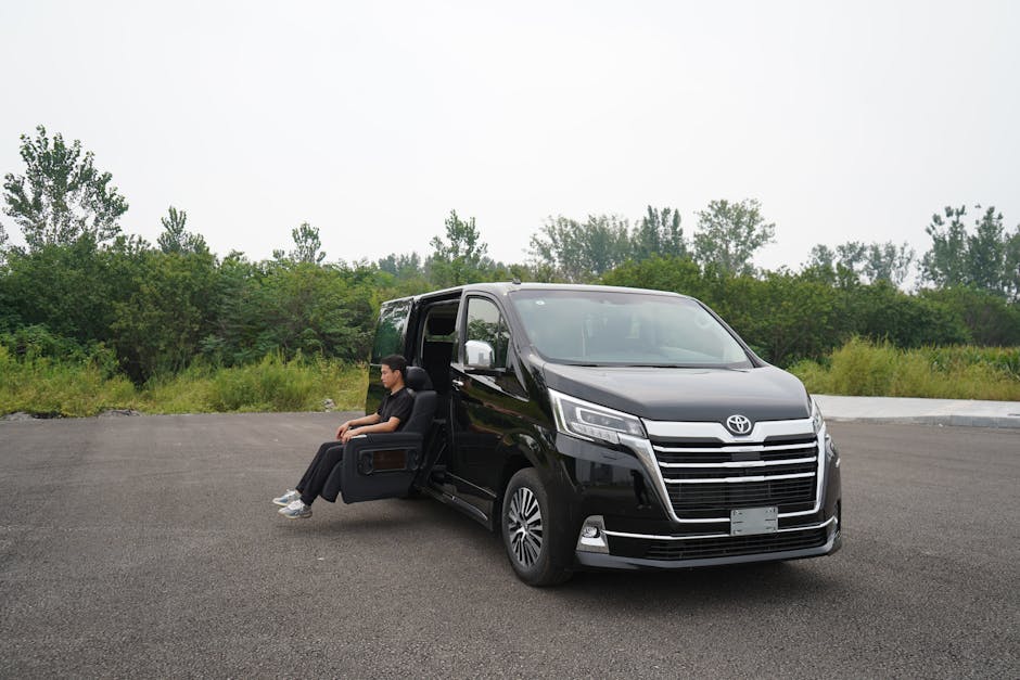 A person sitting in an electric recliner chair positioned next to a black Toyota van parked on an empty paved area, with the individual's legs extending toward the ground. The van's sliding door is open, revealing part of the interior. The scene is outdoors during daytime, with a background of green trees and shrubs under an overcast sky. The setting suggests a loading or unloading process related to home relocation, furniture transport, or moving services, as provided by Man with Van Hainault. The paved surface appears suitable for van parking and access for movers, with no other vehicles or obstructions visible, emphasizing a clean and spacious environment for house removals and packing activities.
