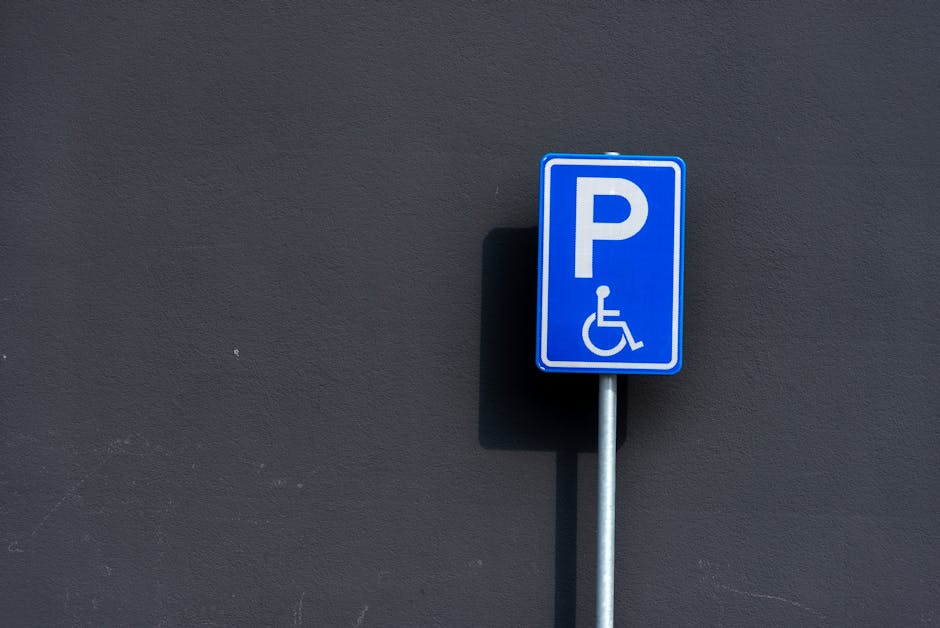 A close-up photograph of a blue parking sign with a white 'P' symbol and a wheelchair accessibility icon, mounted on a silver metal post. The sign is positioned against a smooth, dark gray exterior wall, casting a subtle shadow. The environment appears to be outside a building, with no other objects or vehicles visible in the scene. This parking sign indicates designated disabled parking, relevant for home relocation or furniture transport services provided by Man with Van Hainault, especially when coordinating parking permits during a move day as detailed in the Hainault moving day parking permits guide. The image’s clear lighting and neutral setting support accessibility and SEO focused on house removals and moving logistics.