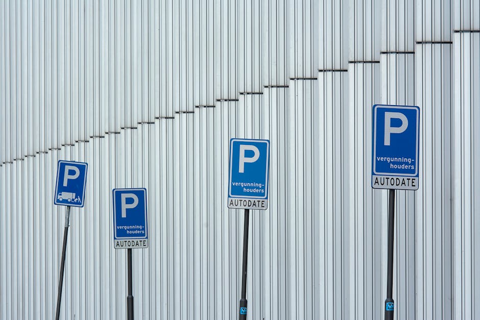 Four blue car parking permit signs mounted on black metal poles in front of a corrugated metal wall. Each sign displays a large white 'P' for parking, with smaller text below indicating 'vergunninghouders' (permit holders) and 'AUTODATE' for automatic renewal. The signs are positioned at varying heights along a paved area, suggesting designated parking zones for residents or authorized vehicles. In the context of house removals and relocation services by Man with Van Hainault, these parking permits relate to parking regulations or permissions required for vehicle access during home relocation or furniture transport. The background features a silver, vertically ridged metal surface, possibly part of a commercial or industrial building, with subtle horizontal lines created by the arrangement of the ridges, under natural lighting conditions.