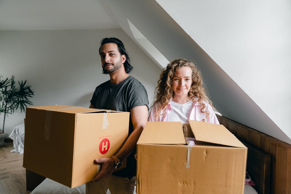 A man and a woman are standing inside a residential property, each holding a large cardboard box sealed with tape. The man, positioned on the left, has dark hair and a beard, wearing a grey t-shirt, and is carrying a box with a red 'H' logo on the side, indicating a professional packing or moving service. The woman, on the right, has curly blonde hair, wearing a white t-shirt with pink accents, and is holding an open cardboard box filled with packing materials such as plastic wrap and paper. They are standing beneath a sloped ceiling with natural light coming from a nearby window or skylight, and a small leafy plant is visible in the background. This scene likely depicts the packing phase of a home relocation, with furniture, boxes, and packing materials prepared for transport by companies like Man with Van Hainault as part of their removals and furniture transport services.