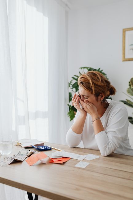 A woman with short, reddish-brown hair, wearing a white long-sleeved top, is seated at a wooden table indoors, with her hands covering her face in an expression of stress or frustration. On the table are various items including a calculator, scattered bills and receipts, envelopes, and some loose papers, indicating financial or moving-related paperwork. Behind her, a large window with sheer white curtains lets natural light into the room, and there are green potted plants and a framed picture on the wall. The scene appears to be a home environment, possibly during a stressful house move or related planning, reflecting the emotional aspect of household relocations that Man with Van Hainault offers as part of their removals services, including packing, moving, and managing relocation costs, as discussed on their website.