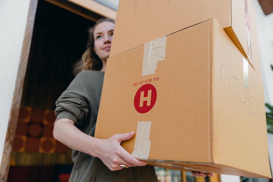A man and a woman are standing inside a residential property, each holding a large cardboard box sealed with tape. The man, positioned on the left, has dark hair and a beard, wearing a grey t-shirt, and is carrying a box with a red 'H' logo on the side, indicating a professional packing or moving service. The woman, on the right, has curly blonde hair, wearing a white t-shirt with pink accents, and is holding an open cardboard box filled with packing materials such as plastic wrap and paper. They are standing beneath a sloped ceiling with natural light coming from a nearby window or skylight, and a small leafy plant is visible in the background. This scene likely depicts the packing phase of a home relocation, with furniture, boxes, and packing materials prepared for transport by companies like Man with Van Hainault as part of their removals and furniture transport services.