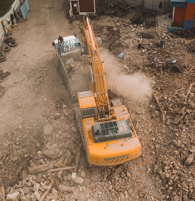 An aerial view of a large yellow demolition excavator operating on a construction site surrounded by dirt, rubble, and debris. The machine is positioned over a partially demolished area with broken br