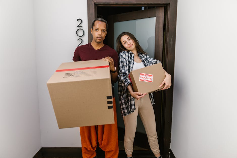 A man and a woman are standing in a narrow hallway beside an open doorway, each holding a packed cardboard box. The man, wearing a maroon T-shirt and orange trousers, holds a large box with a red tape seal, while the woman, dressed in a checkered shirt and beige pants, carries a smaller box labeled 'Fragile.' Both are positioned on a concrete or tiled floor, with the man standing slightly to the left and the woman to the right. The background includes a white wall with the number '200' displayed vertically and a dark-colored door frame leading into another room. Visible behind the doorway is a blue wall, suggesting an interior home environment during a house move or relocation. The scene depicts the process of packing or unpacking for a home relocation, in line with furniture transport and moving logistics, as facilitated by Man with Van Hainault, a professional removals service.