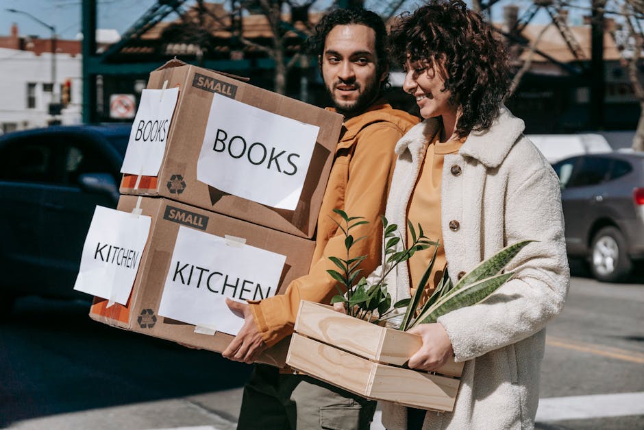Two individuals are participating in a house move outside a residential property, with one person carrying two large cardboard boxes labelled 'BOOKS' and 'KITCHEN' in black text, used for packing household items. The boxes are secured with packing tape and are held close to their chest as they walk along a pavement, with parked cars and a building with a wooden roof visible in the background. Beside them, the other person holds a wooden crate containing small potted plants with green foliage, representing packing of household or decorative items. The scene is set outdoors during daylight, with natural light illuminating the participants and their surroundings. The moving process involves careful handling of packed possessions, aligning with house removal and furniture transport activities, and giving a clear visual context for home relocation tasks with a focus on packing and loading procedures, as managed by a professional removals service like Man with Van Hainault.