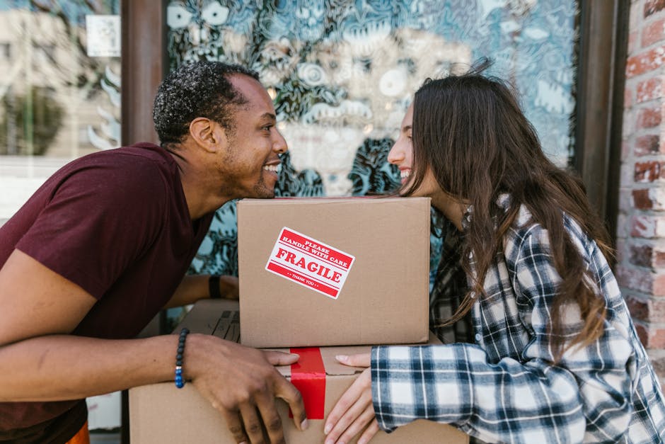 A man and a woman are standing in a narrow hallway beside an open doorway, each holding a packed cardboard box. The man, wearing a maroon T-shirt and orange trousers, holds a large box with a red tape seal, while the woman, dressed in a checkered shirt and beige pants, carries a smaller box labeled 'Fragile.' Both are positioned on a concrete or tiled floor, with the man standing slightly to the left and the woman to the right. The background includes a white wall with the number '200' displayed vertically and a dark-colored door frame leading into another room. Visible behind the doorway is a blue wall, suggesting an interior home environment during a house move or relocation. The scene depicts the process of packing or unpacking for a home relocation, in line with furniture transport and moving logistics, as facilitated by Man with Van Hainault, a professional removals service.