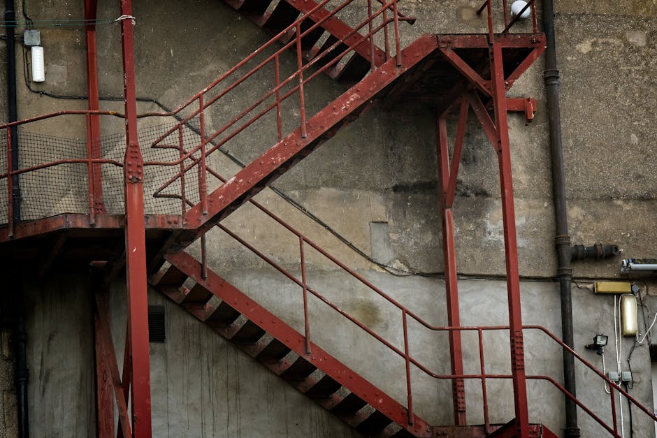 A red metal fire escape staircase attached to an exterior concrete wall of a building, with several landings and staircases connecting at different levels. The staircase features rusted metal railings, mesh panels on the upper platform, and metal steps with open risers. The surrounding wall shows signs of weathering and staining, with visible pipes and electrical components mounted on the wall near the staircase. The stairs are positioned adjacent to a doorway leading into a residential or commercial property, with the staircase providing access via external entry points. This image captures a typical urban fire escape structure that may be encountered during the home relocation process, especially when navigating staircases for furniture transport in buildings with multiple floors during house removals managed by Man with Van Hainault.