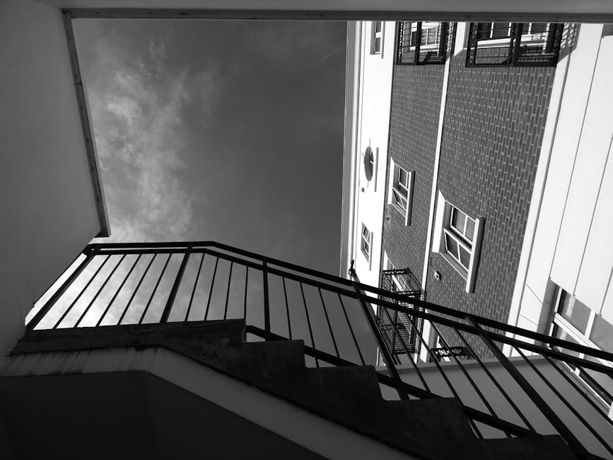 A red metal fire escape staircase attached to an exterior concrete wall of a building, with several landings and staircases connecting at different levels. The staircase features rusted metal railings, mesh panels on the upper platform, and metal steps with open risers. The surrounding wall shows signs of weathering and staining, with visible pipes and electrical components mounted on the wall near the staircase. The stairs are positioned adjacent to a doorway leading into a residential or commercial property, with the staircase providing access via external entry points. This image captures a typical urban fire escape structure that may be encountered during the home relocation process, especially when navigating staircases for furniture transport in buildings with multiple floors during house removals managed by Man with Van Hainault.