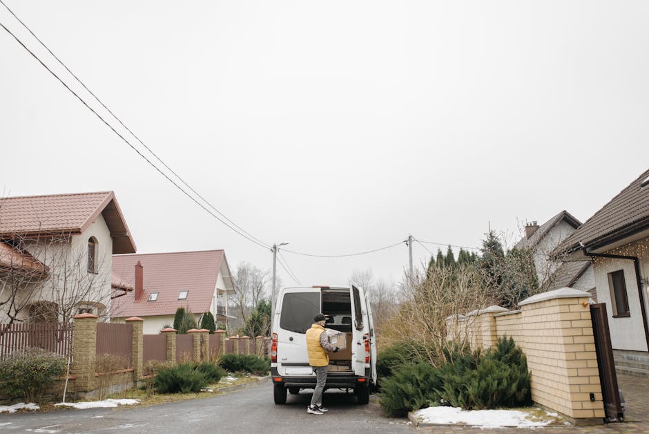 A man wearing a yellow jacket and a black hat is loading a cardboard box into the back of a white cargo van parked on a residential street. The van is positioned on the pavement in front of a house with a beige brick wall and a brown gate, amidst a suburban neighbourhood with other homes featuring sloped tiled roofs. Surrounding the scene are bushes, some of which have patches of snow nearby on the ground. Overhead, electrical wires run across the overcast sky. The image captures the process of home relocation, with the van used for furniture transport and packing and moving activities associated with house removals. Man with Van Hainault occasionally provides such services, including loading and unloading during the moving process.
