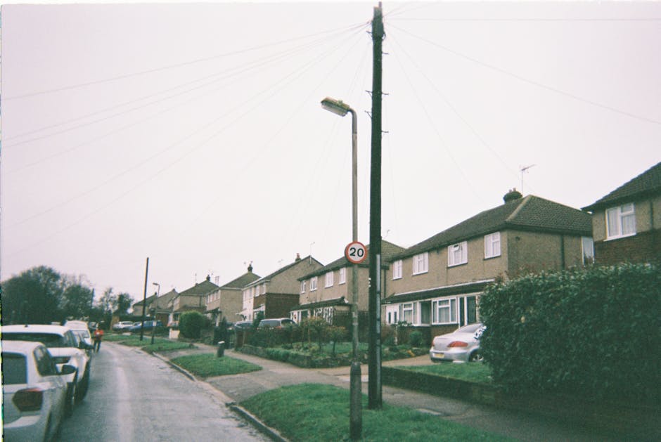 A residential street scene in Hainault, featuring a row of semi-detached houses with front gardens and parked cars along the pavement. In the foreground, a black utility pole with street lighting and a 20 mph speed limit sign is visible, with overhead power lines extending across the sky. The street appears wet, indicating recent rain, with overcast weather casting diffuse lighting. The houses are two-storey, built with brick and siding, and have small front yards bordered by low hedges or fences. Some driveway areas are visible, though there is no driveway in this particular location, which may affect home relocation logistics. Occasional parked vehicles include a silver sedan and a white van, with a narrow, paved sidewalk running along the edge of the street. These elements illustrate a typical UK suburban environment that [COMPANY_NAME], such as Man with Van Hainault, could assist with during packing and furniture transport as part of house removals or moving services.