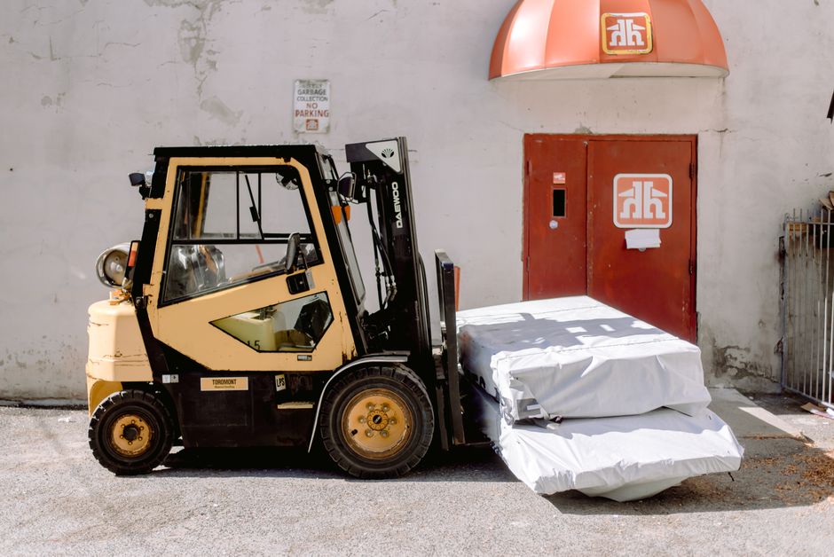 A man wearing a yellow jacket and a black hat is loading a cardboard box into the back of a white cargo van parked on a residential street. The van is positioned on the pavement in front of a house with a beige brick wall and a brown gate, amidst a suburban neighbourhood with other homes featuring sloped tiled roofs. Surrounding the scene are bushes, some of which have patches of snow nearby on the ground. Overhead, electrical wires run across the overcast sky. The image captures the process of home relocation, with the van used for furniture transport and packing and moving activities associated with house removals. Man with Van Hainault occasionally provides such services, including loading and unloading during the moving process.