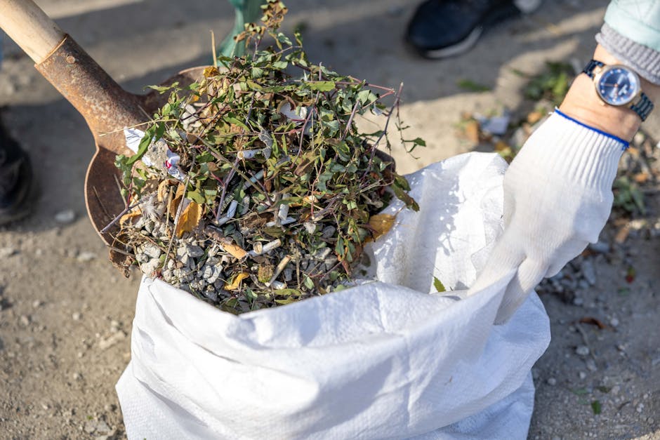 A person wearing a white glove and a wristwatch is holding a white sack filled with garden debris, including small branches, leaves, and twigs, during a home removal or clearance process. The ground is composed of gravel and dirt, with some scattered plant matter visible. A metal shovel with a wooden handle is positioned in the sack, indicating active removal or loading of garden waste, possibly for recycling or disposal as part of house removals or local waste regulations in Hainault. The scene is outdoors, and the individual’s arm suggests they are engaged in packing or transporting waste materials for a house relocation service offered by Man with Van Hainault, supporting the process of packing and moving related to household clearance activities.