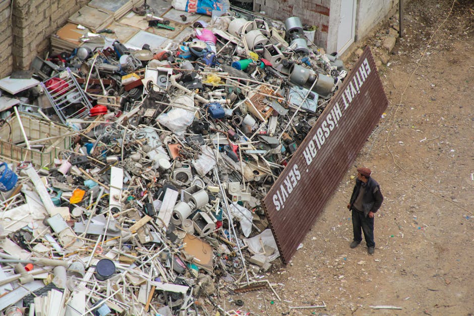 An aerial view of a large pile of household waste and discarded items, including broken appliances, plastic containers, and metal scraps, spread across a gravel surface next to a brick wall. A brown metal sign with white lettering reading 'HAILSHAM BULKY WASTE' is positioned at an angle, partially obscuring part of the debris. Nearby, a man dressed in dark clothing and a brown hat stands on the dirt ground, observing the clutter. The scene is outdoors with natural lighting, illustrating a waste collection or disposal site adjacent to a residential area. The presence of this waste pile highlights the importance of proper house removals and waste management services offered by Man with Van Hainault, especially when preparing for a home relocation or furniture transport. The environment emphasizes the need for organized clearing and adherence to local waste disposal rules during house moves in the Hainault area.