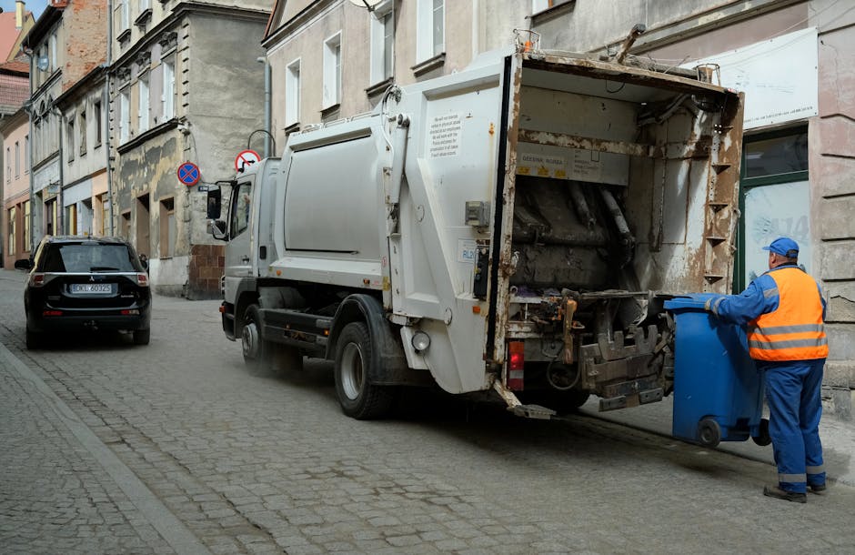 A person wearing a white glove and a wristwatch is holding a white sack filled with garden debris, including small branches, leaves, and twigs, during a home removal or clearance process. The ground is composed of gravel and dirt, with some scattered plant matter visible. A metal shovel with a wooden handle is positioned in the sack, indicating active removal or loading of garden waste, possibly for recycling or disposal as part of house removals or local waste regulations in Hainault. The scene is outdoors, and the individual’s arm suggests they are engaged in packing or transporting waste materials for a house relocation service offered by Man with Van Hainault, supporting the process of packing and moving related to household clearance activities.