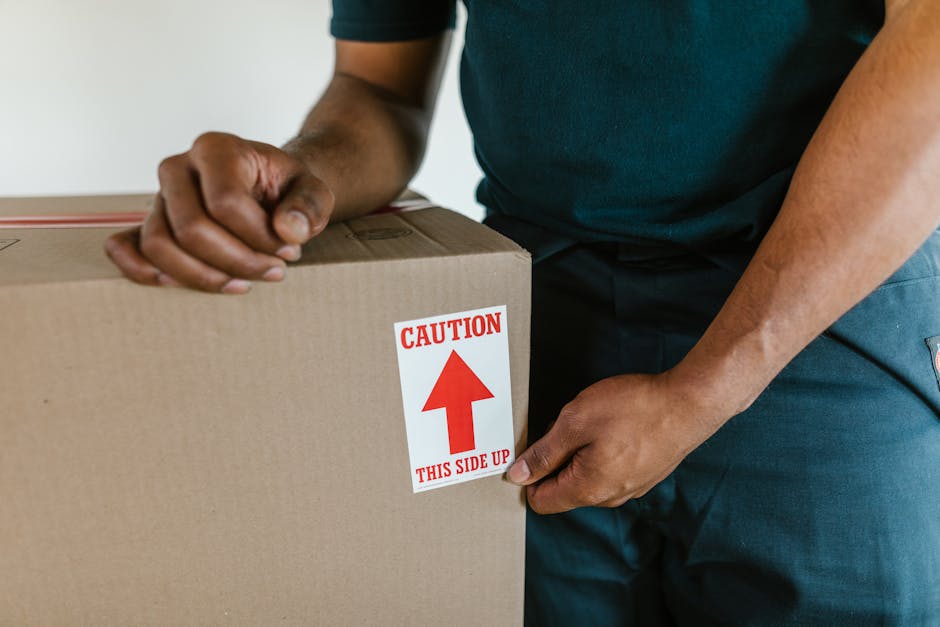 Close-up view of a person in casual clothing, holding a large cardboard box with both hands during a home relocation. The box features a red and white caution label with an upward arrow and the instruction 'THIS SIDE UP,' indicating proper orientation. The individual is inside a property, standing near a doorway or open space, with the background showing a neutral-colored wall. The box appears to be made of sturdy cardboard and is likely packed with household items for transport. Visible parts of the person's arms and hands suggest careful handling during the packing and loading process. The image captures the meticulous nature of furniture transport and packing involved in residential removals, reflecting the tasks undertaken by companies like Man with Van Hainault as part of their essential move-day procedures.
