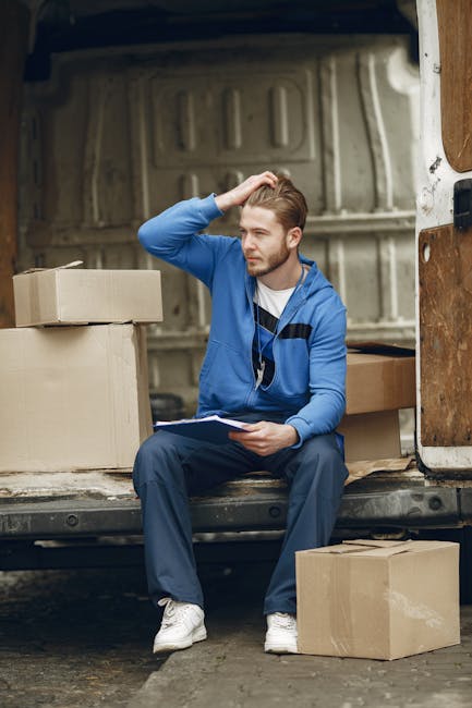 A man wearing a white polo shirt and black cap is standing outside next to a moving van during a house relocation. He is smiling and holding a medium-sized cardboard box, ready for loading. The back o