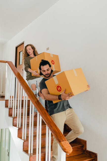 A man and a woman are carrying cardboard boxes as part of a home relocation process inside a residential property. The man, with dark hair and a beard, is descending wooden stairs while holding a large box wrapped with packing tape and marked with a red symbol. The woman, standing behind him on the staircase, is holding a similar box and is dressed in a casual top and ripped jeans. The staircase has a polished wooden handrail and white spindles, with the interior walls painted in a neutral light shade. The scene suggests an organized packing and moving operation, likely conducted by a professional removals service such as Man with Van Hainault, involved in furniture transport and packing logistics for quick, same-day house removals.